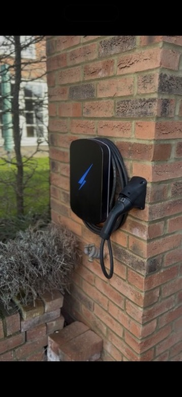 Black Hypervolt EV charger with a coiled tethered cable installed on a red brick pillar next to a garden